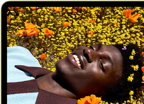 MacBook Neo, Farbe Zitrus, das Display zeigt eine Person, die in einer Blumenwiese in der Sonne liegt und lächelt