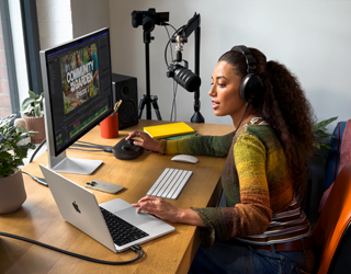 Eine Frau arbeitet bei sich zu Hause an ihrem Schreibtisch mit einem MacBook Pro, das an zwei externe Displays angeschlossen ist. Sie trägt Kopfhörer
